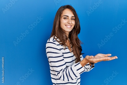 Young hispanic woman standing over blue isolated background pointing aside with hands open palms showing copy space, presenting advertisement smiling excited happy © Krakenimages.com