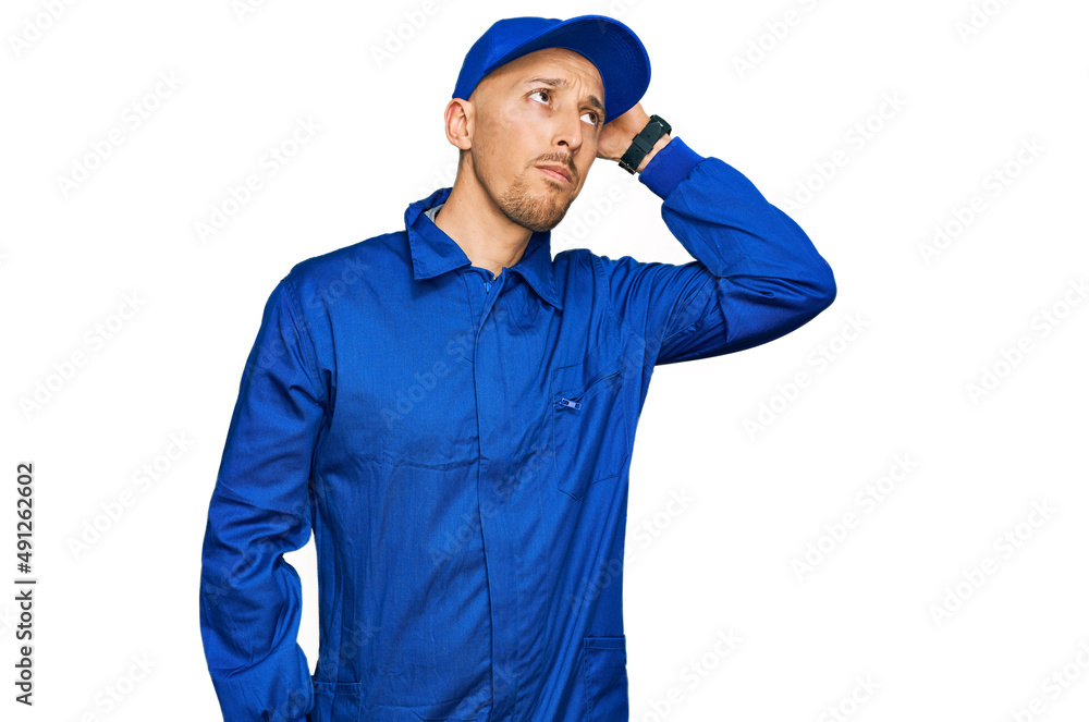 Bald man with beard wearing builder jumpsuit uniform confuse and wondering about question. uncertain with doubt, thinking with hand on head. pensive concept.