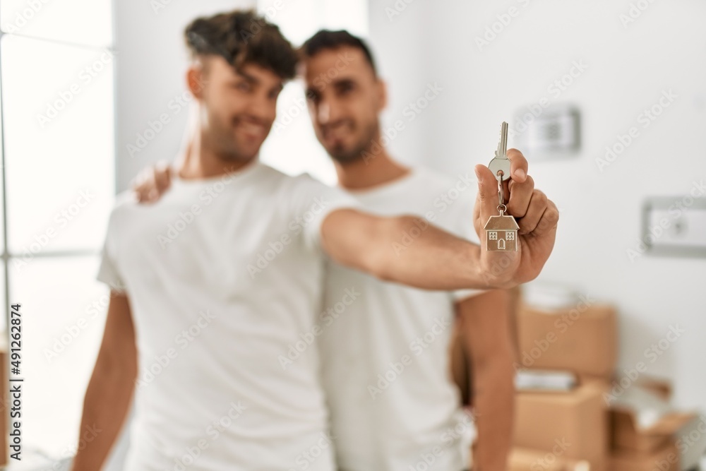 Two hispanic men couple hugging each other holding key of new house at new home Two hispanic men couple hugging each other holding key of new house at new home