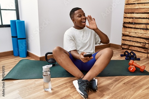 Young african man sitting on training mat at the gym using smartphone shouting and screaming loud to side with hand on mouth. communication concept.