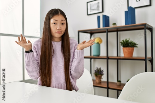 Young chinese girl wearing casual clothes sitting on the table at home clueless and confused expression with arms and hands raised. doubt concept.