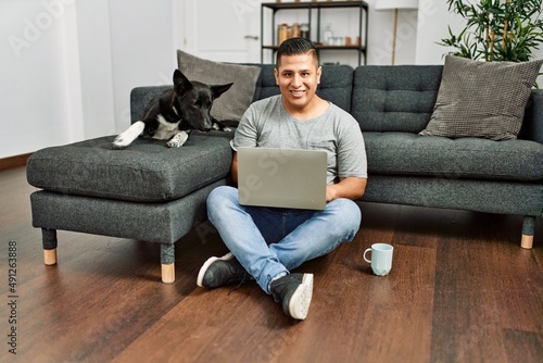 Young hispanic man using laptop sitting on the floor with dog at home. © Krakenimages.com
