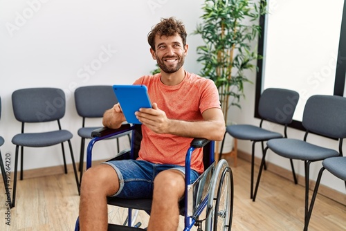 Young hispanic man using touchpad sitting on wheelchair at clinic waiting room.