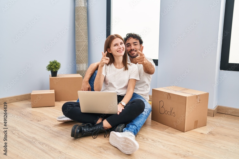 Young couple using laptop at new home pointing fingers to camera with happy and funny face. good energy and vibes. Young couple using laptop at new home pointing fingers to camera with happy and funny face. good energy and vibes.