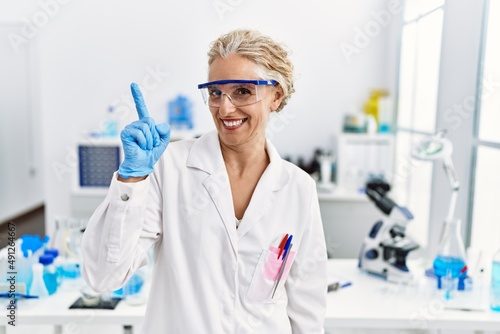 Middle age blonde woman working at scientist laboratory pointing finger up with successful idea. exited and happy. number one.
