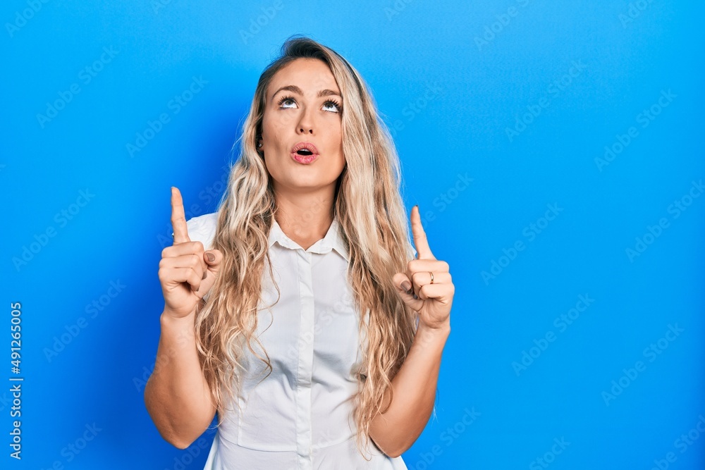 Fototapeta premium Beautiful young blonde woman wearing casual white shirt amazed and surprised looking up and pointing with fingers and raised arms.