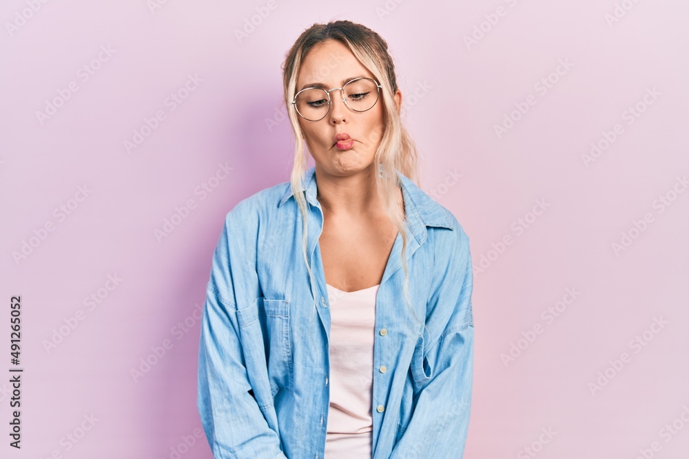 Beautiful young blonde woman wearing casual clothes and glasses making fish face with lips, crazy and comical gesture. funny expression.