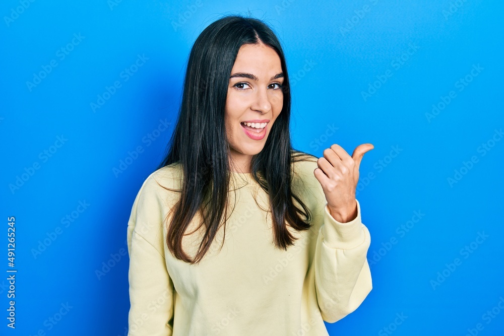 Young brunette woman wearing casual sweatshirt smiling with happy face looking and pointing to the side with thumb up. Young brunette woman wearing casual sweatshirt smiling with happy face looking and pointing to the side with thumb up.