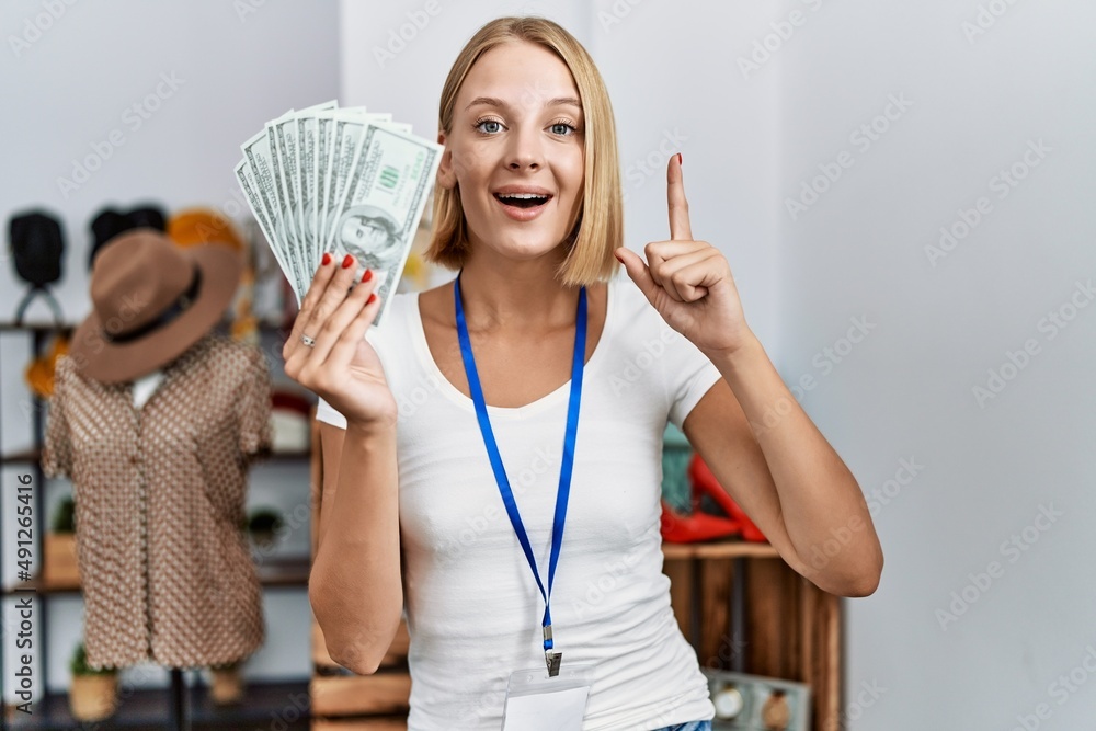 Young caucasian woman working at retail boutique holding dollars banknotes smiling with an idea or question pointing finger with happy face, number one Young caucasian woman working at retail boutique holding dollars banknotes smiling with an idea or question pointing finger with happy face, number one