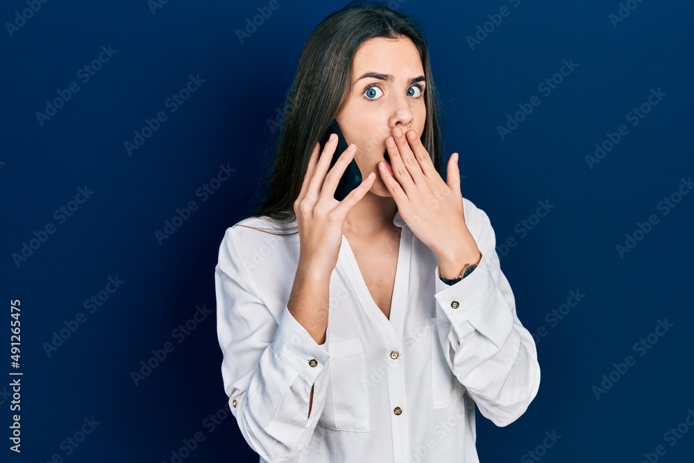 Young brunette teenager having conversation talking on the smartphone covering mouth with hand, shocked and afraid for mistake. surprised expression Young brunette teenager having conversation talking on the smartphone covering mouth with hand, shocked and afraid for mistake. surprised expression