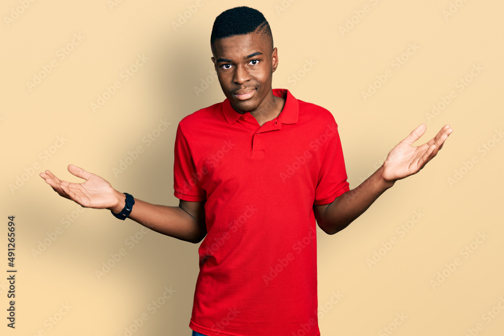 Young african american man wearing casual red t shirt clueless and confused expression with arms and hands raised. doubt concept. Young african american man wearing casual red t shirt clueless and confused expression with arms and hands raised. doubt concept.