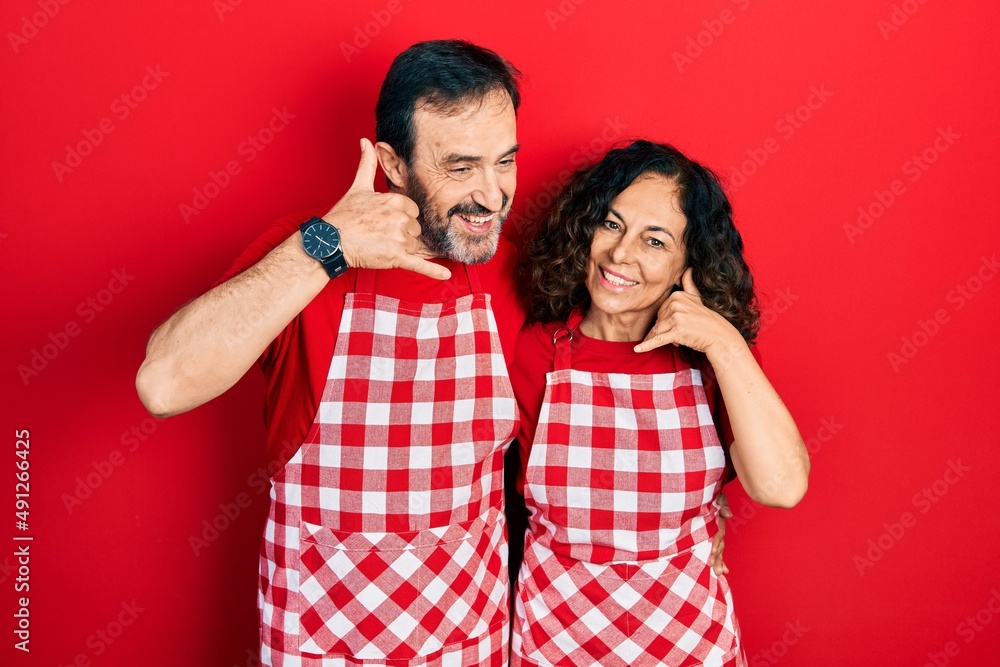 Middle age couple of hispanic woman and man wearing cook apron smiling doing phone gesture with hand and fingers like talking on the telephone. communicating concepts. Middle age couple of hispanic woman and man wearing cook apron smiling doing phone gesture with hand and fingers like talking on the telephone. communicating concepts.