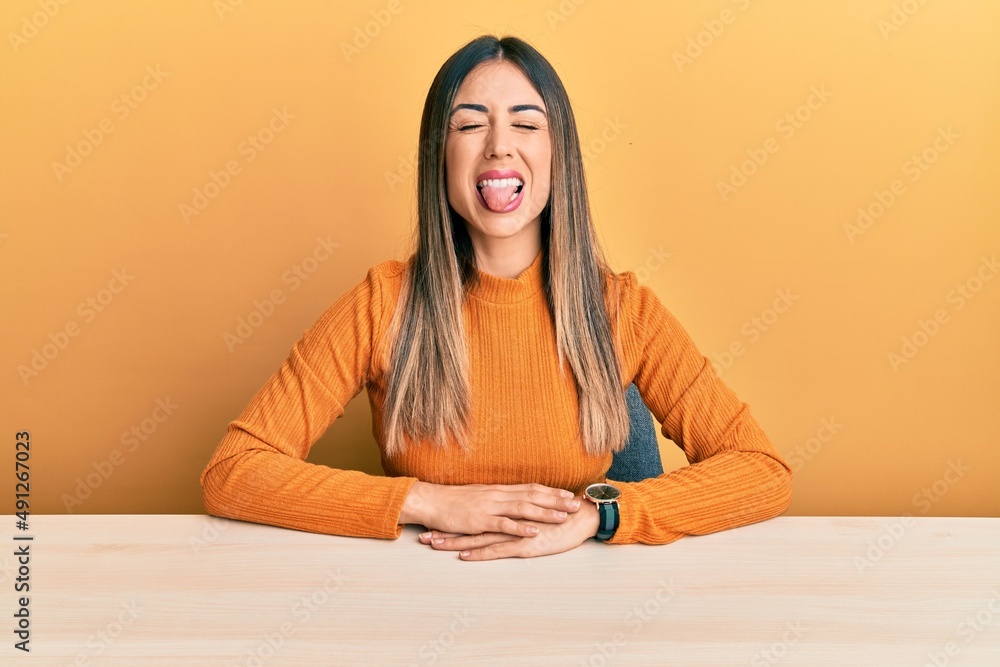Young hispanic woman wearing casual clothes sitting on the table sticking tongue out happy with funny expression. emotion concept. Young hispanic woman wearing casual clothes sitting on the table sticking tongue out happy with funny expression. emotion concept.