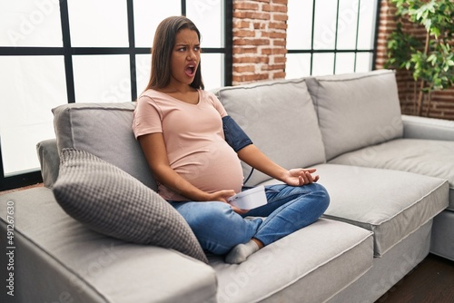 Young pregnant woman using blood pressure monitor sitting on the sofa angry and mad screaming frustrated and furious, shouting with anger. rage and aggressive concept.