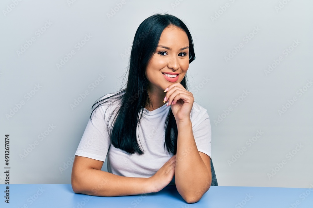 Beautiful hispanic woman with nose piercing wearing casual white t shirt sitting on the table smiling looking confident at the camera with crossed arms and hand on chin. thinking positive. Beautiful hispanic woman with nose piercing wearing casual white t shirt sitting on the table smiling looking confident at the camera with crossed arms and hand on chin. thinking positive.