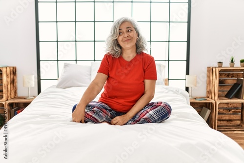 Middle age woman with grey hair sitting on the bed at home looking away to side with smile on face, natural expression. laughing confident.