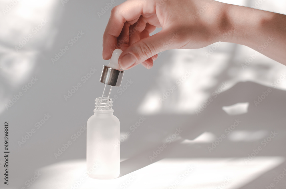 Female hand holding pipette with essential oil or serum. Natural cosmetics for body care. White cosmetic bottle and female hand on a white background with shadows