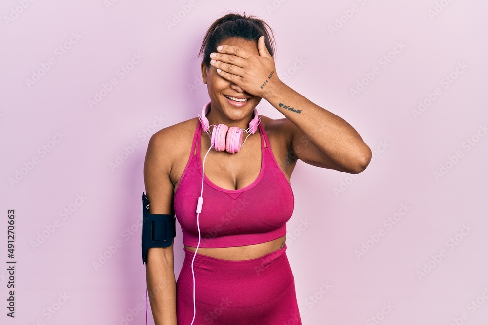 Young african american girl wearing gym clothes and using headphones smiling and laughing with hand on face covering eyes for surprise. blind concept.