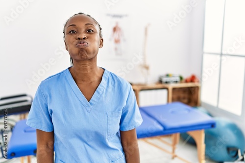 Black woman with braids working at pain recovery clinic puffing cheeks with funny face. mouth inflated with air  crazy expression.