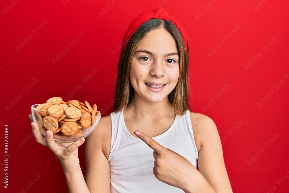 Beautiful brunette little girl holding salty biscuits in a bowl smiling happy pointing with hand and finger Beautiful brunette little girl holding salty biscuits in a bowl smiling happy pointing with hand and finger