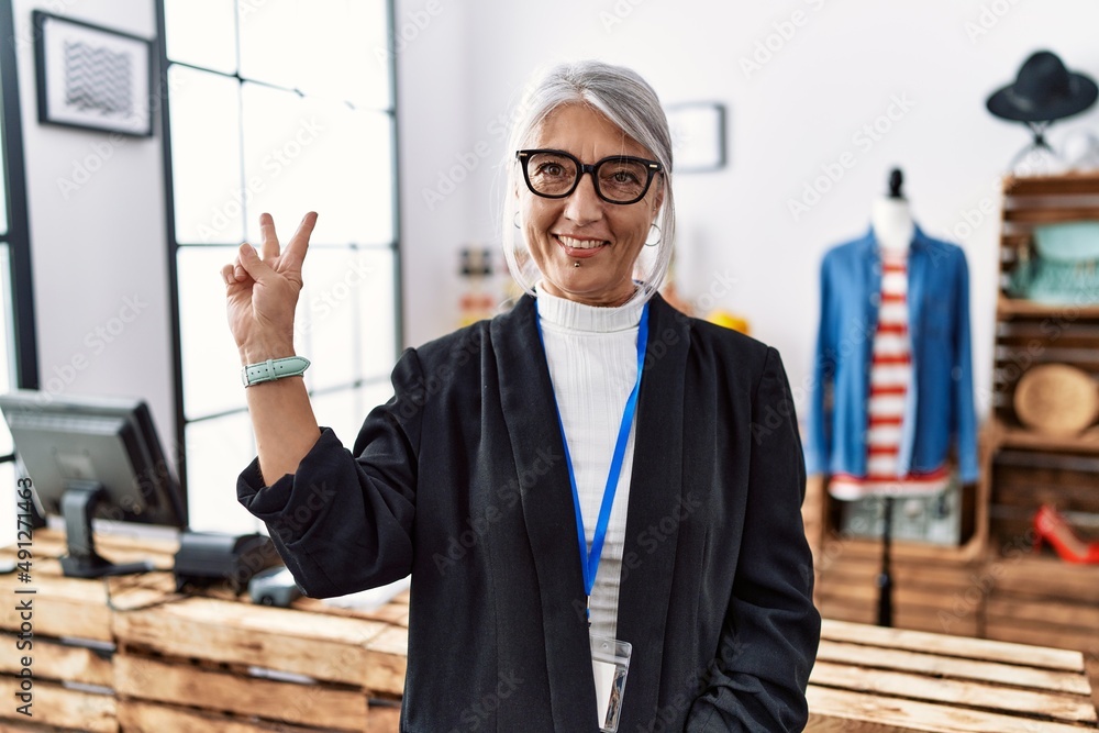 Middle age grey-haired woman working as manager at retail boutique smiling with happy face winking at the camera doing victory sign. number two. Middle age grey-haired woman working as manager at retail boutique smiling with happy face winking at the camera doing victory sign. number two.