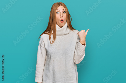 Young irish woman wearing casual winter sweater surprised pointing with hand finger to the side, open mouth amazed expression.