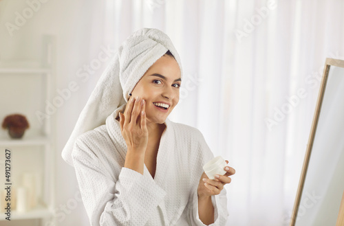 Portrait of beautiful young woman taking care of her skin on her face while standing in bathroom. Girl in bathrobe and with towel on her head holds container of skin cream and smears it on her face.