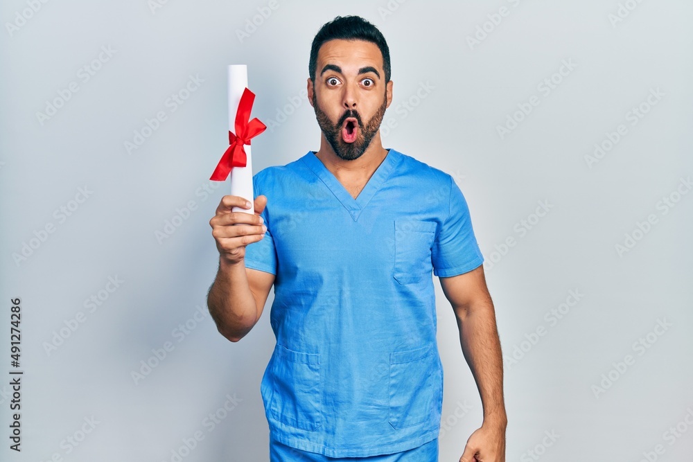 Handsome hispanic man with beard wearing blue male nurse uniform holding diploma scared and amazed with open mouth for surprise, disbelief face Handsome hispanic man with beard wearing blue male nurse uniform holding diploma scared and amazed with open mouth for surprise, disbelief face