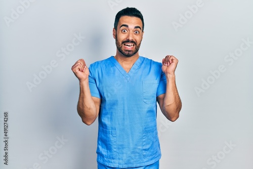 Handsome hispanic man with beard wearing blue male nurse uniform celebrating surprised and amazed for success with arms raised and open eyes. winner concept.