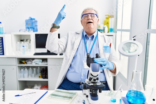Senior caucasian man working at scientist laboratory smiling amazed and surprised and pointing up with fingers and raised arms.
