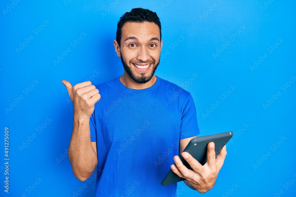 Hispanic man with beard using touchpad device pointing thumb up to the side smiling happy with open mouth Hispanic man with beard using touchpad device pointing thumb up to the side smiling happy with open mouth