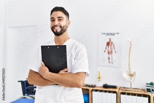 Young arab man wearing physiotherapist uniform holding clipboard at clinic