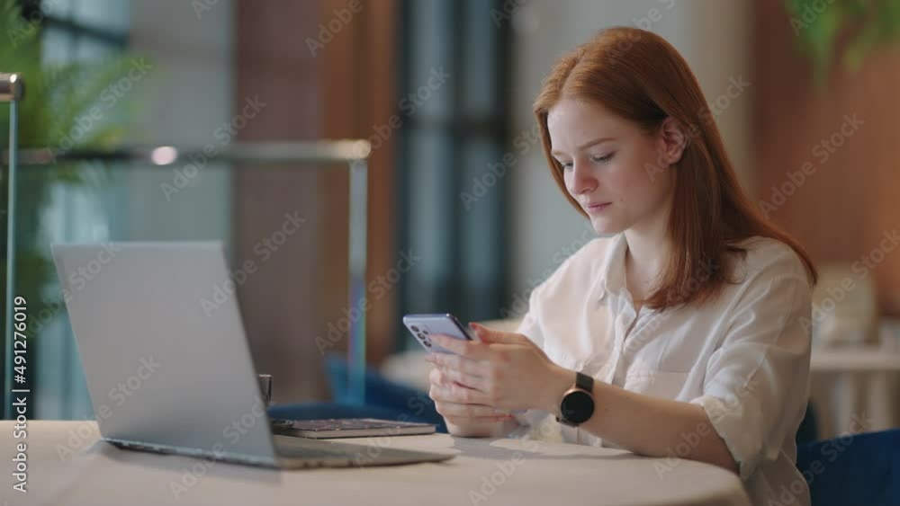 Red-haired Woman working from home using laptop computer while reading text message on mobile phone. Woman using a phone. Serious charming woman using smartphone while working with laptop at home Red-haired Woman working from home using laptop computer while reading text message on mobile phone. Woman using a phone. Serious charming woman using smartphone while working with laptop at home