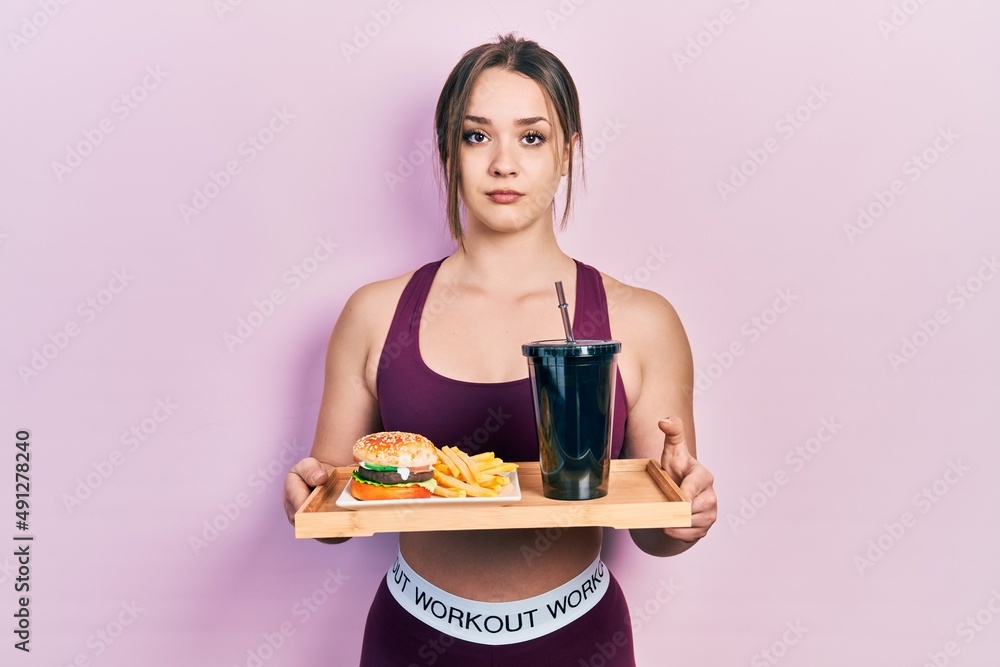 Young hispanic girl wearing sportswear eating a tasty classic burger with fries and soda relaxed with serious expression on face. simple and natural looking at the camera. Young hispanic girl wearing sportswear eating a tasty classic burger with fries and soda relaxed with serious expression on face. simple and natural looking at the camera.