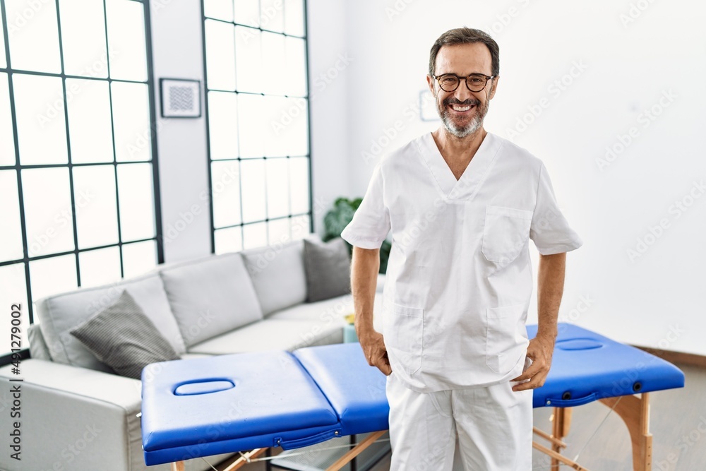 Middle age physiotherapy man working at home looking positive and happy standing and smiling with a confident smile showing teeth Middle age physiotherapy man working at home looking positive and happy standing and smiling with a confident smile showing teeth