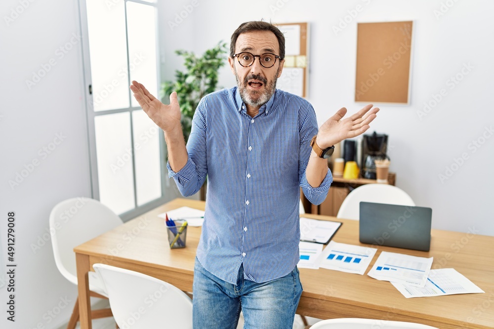 Middle age hispanic man with beard wearing business clothes at the office clueless and confused expression with arms and hands raised. doubt concept. Middle age hispanic man with beard wearing business clothes at the office clueless and confused expression with arms and hands raised. doubt concept.
