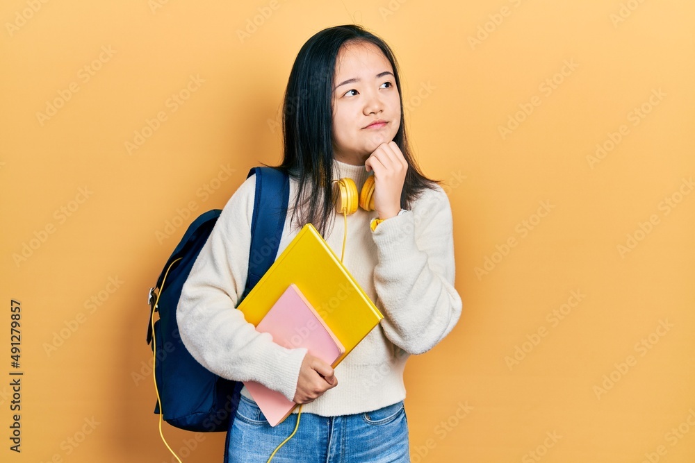 Young chinese girl holding student backpack and books with hand on chin thinking about question, pensive expression. smiling with thoughtful face. doubt concept. Young chinese girl holding student backpack and books with hand on chin thinking about question, pensive expression. smiling with thoughtful face. doubt concept.