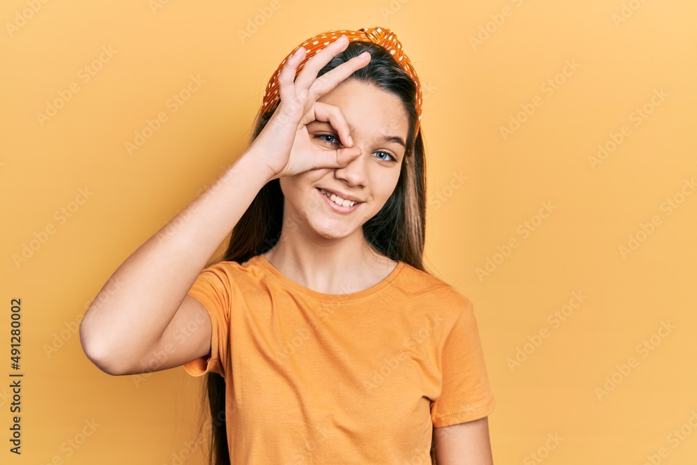 Young brunette girl wearing casual orange t shirt smiling happy doing ok sign with hand on eye looking through fingers