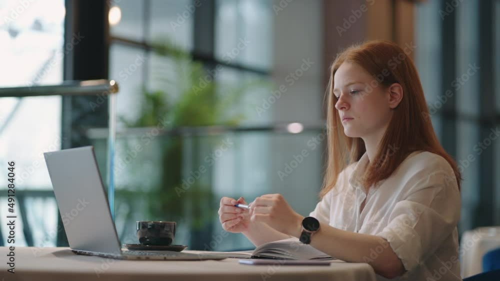 thoughtful brooding remote working red haired woman sitting infront of a laptop or notebook in casual outfit on her work desk in her modern airy bright living room home office with many windows thoughtful brooding remote working red haired woman sitting infront of a laptop or notebook in casual outfit on her work desk in her modern airy bright living room home office with many windows