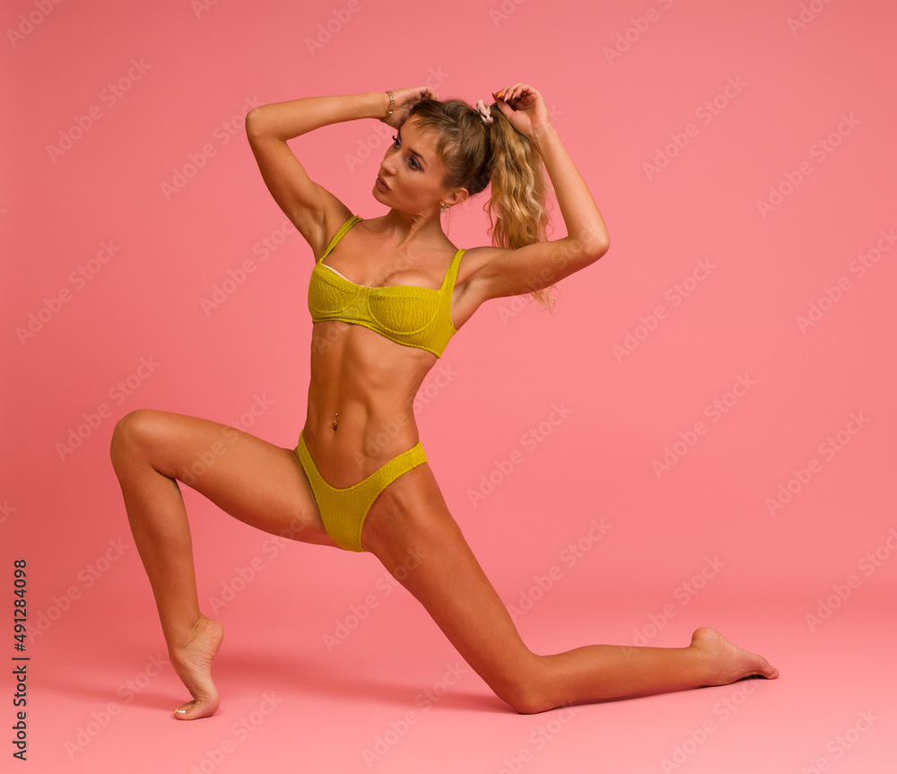 Slender sexy woman in a yellow swimsuit posing while standing on one knee on a pink background.