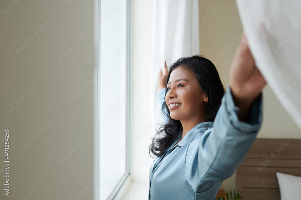 Happy young woman in silk loungewear opening curtains and looking through window enjoying new morning Happy young woman in silk loungewear opening curtains and looking through window enjoying new morning