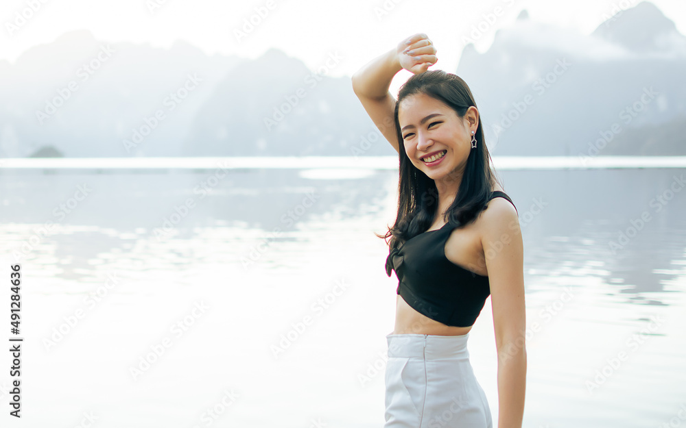 Selective focus woman wearing casual clothes, standing beside lake or river with blur mountains, taking rest, raising hands with relaxing, refreshment, happiness. Summer Travel in Holiday Concept. Selective focus woman wearing casual clothes, standing beside lake or river with blur mountains, taking rest, raising hands with relaxing, refreshment, happiness. Summer Travel in Holiday Concept.