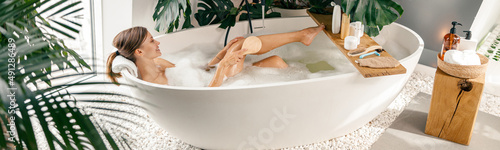 Canvas-taulu Relaxed young woman using brush while taking buble bath in modern bathroom decorated with tropical plants
