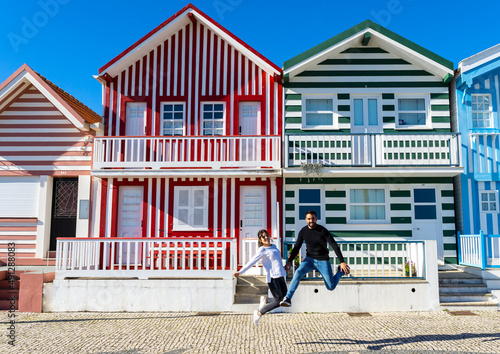 Woman and man jumping for a photo in front of the typical colorful striped houses in Costa Nova, Portugal.