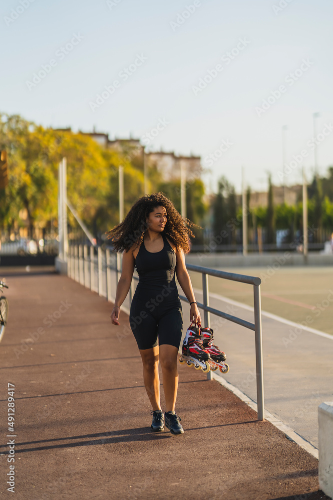 Chica marroquí con pelo largo rizado andando con patines en la mano por espacio deportivo Chica marroquí con pelo largo rizado andando con patines en la mano por espacio deportivo
