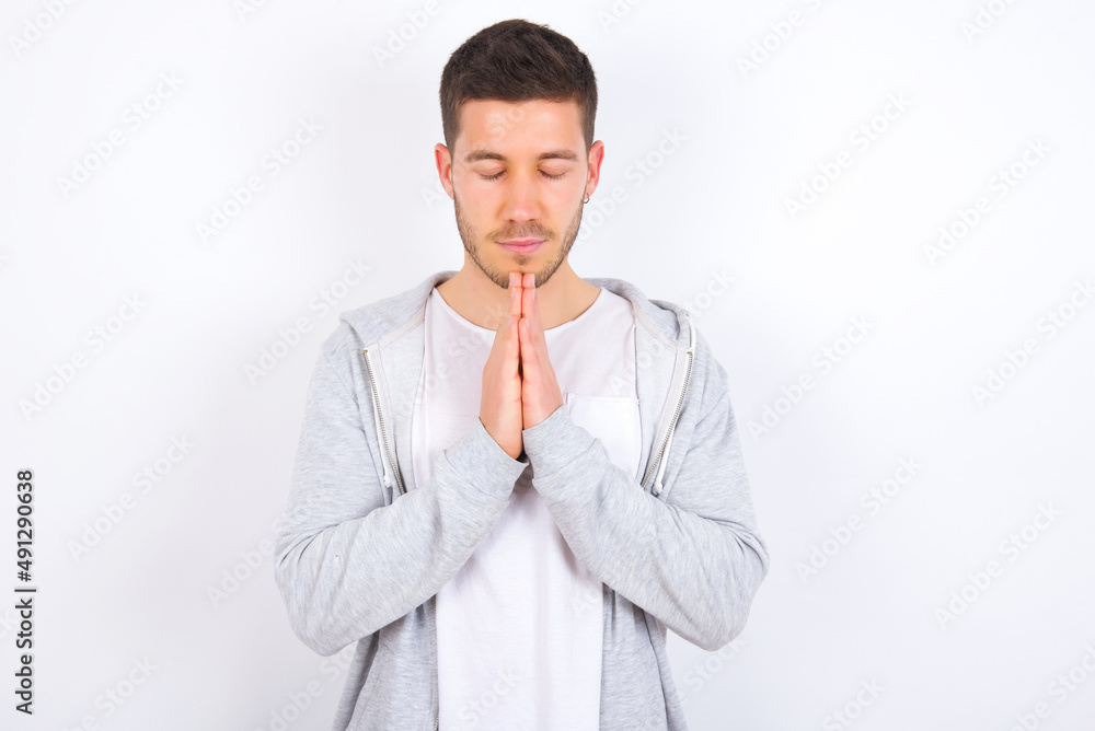 Indoor closeup of young caucasian man wearing casual clothes over white background practicing yoga and meditation, holding palms together in namaste, looking calm, relaxed and peaceful. Indoor closeup of young caucasian man wearing casual clothes over white background practicing yoga and meditation, holding palms together in namaste, looking calm, relaxed and peaceful.
