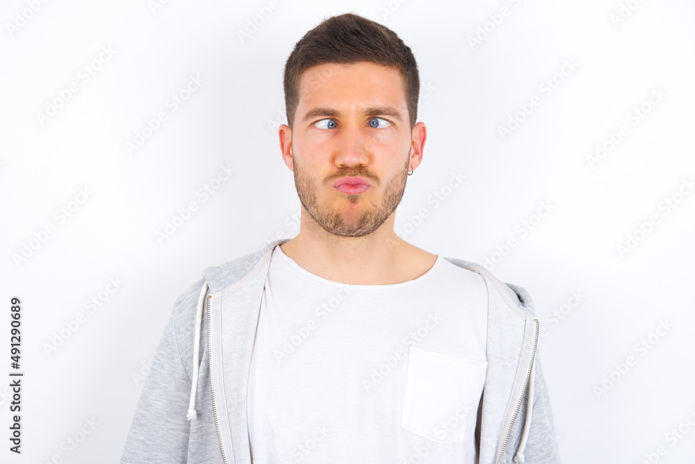 young caucasian man wearing casual clothes over white background crosses eyes, puts lips, makes grimace with awkward expression has fun alone, plays fool. young caucasian man wearing casual clothes over white background crosses eyes, puts lips, makes grimace with awkward expression has fun alone, plays fool.