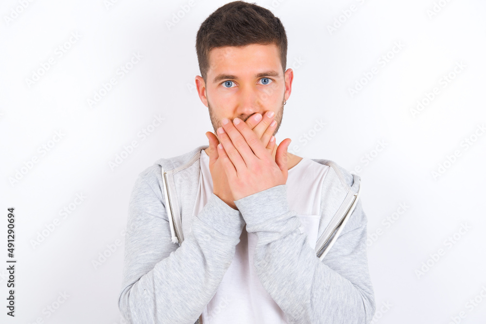 Upset young caucasian man wearing casual clothes over white background, covering her mouth with both palms to prevent screaming sound, after seeing or hearing something bad. Upset young caucasian man wearing casual clothes over white background, covering her mouth with both palms to prevent screaming sound, after seeing or hearing something bad.
