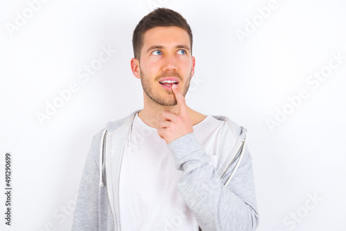 young caucasian man wearing casual clothes over white background with thoughtful expression, looks to the camera, keeps hand near face, bitting a finger thinks about something pleasant.