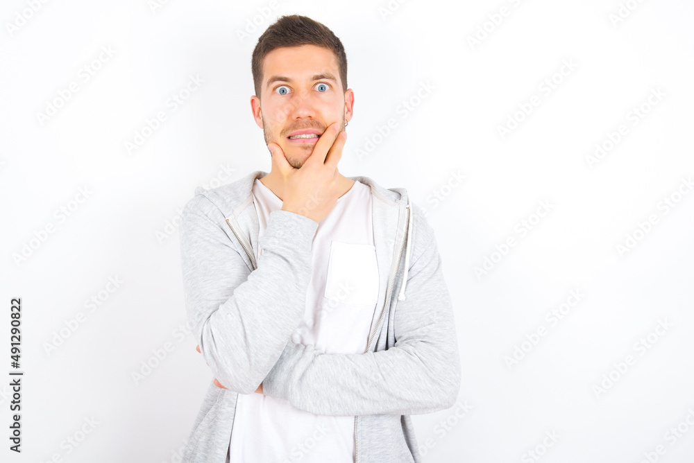 young caucasian man wearing casual clothes over white background covering mouth with hands scared from something or someone bitting nails young caucasian man wearing casual clothes over white background covering mouth with hands scared from something or someone bitting nails